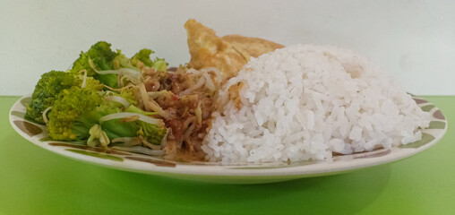 Side view food menu is pecel consisting of white rice, broccoli, sprouts, fried tempe, fried egg and pecel seasoning in the green table.
