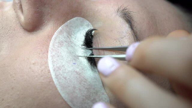 Skillful cosmetologist increases eyelashes with a volumetric method in a beauty salon. The process of increasing and volumetric eyelash extensions to a girl. Gluing false lashes with a tweezers.