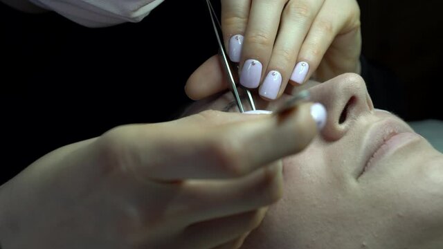 Closeup of eyelash extension procedure. Woman eye with long curvy eyelashes. Young woman undergoing eyelash extensions procedure, closeup. Gluing false lashes with a tweezers.