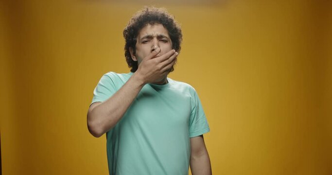 Young asian man with curly hair in casual clothes beind tired or bored, yawning and covering his mouth, isolated on yellow background close up 4k