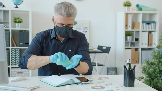 Man in reusable face mask and gloves is wet wiping hands with sanitizer in modern office concerned with hygiene during infectious disease epidemic.