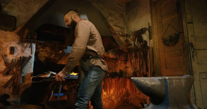 Young handsome male smith with long beard taking out molten glowing metal horseshoe from fire flame and looking at it with interest in antique smithy. Brutal forger working in workshop manufactory.