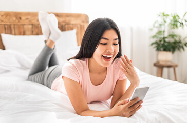 Excited Asian Girl With Cellphone Reading Message Lying On Bed