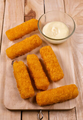 Pile of golden fried fish fingers with white garlic sauce placed on chopping board on wooden background