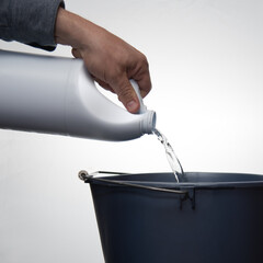 A man's hand pouring cleaner or bleach into a blue bucket against a white background.