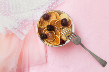 Mini pancakes with blackberry white dishes on a pink table. Lovely breakfast, morning light. A new trend dessert in social networks. Little Dutch poffertjes. Top view, flat lay.