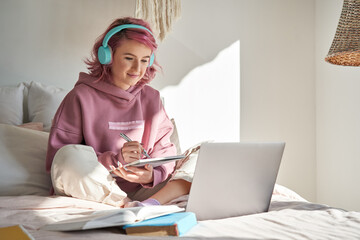 Fototapeta Hipster teen girl school student with pink hair wear headphone write notes watch video online webinar learn on laptop sit in bed distance elearning course video conference pc call in bedroom at home. obraz