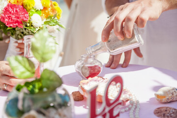 Bride and groom pouring colorful different colored sands into the crystal vase close up during symbolic nautical decor destination wedding marriage unity ceremony on sandy beach in front of the ocean 