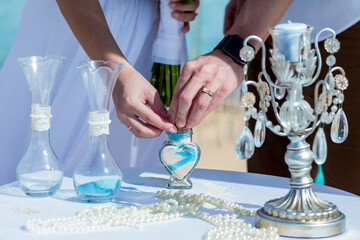 Bride and groom pouring colorful different colored sands into the crystal vase close up during symbolic nautical decor destination wedding marriage unity ceremony on sandy beach in front of the ocean 