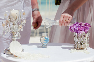 Bride and groom pouring colorful different colored sands into the crystal vase close up during symbolic nautical decor destination wedding marriage unity ceremony on sandy beach in front of the ocean 