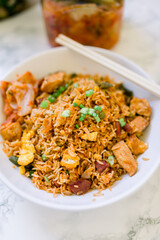 Delicious Chicken Fried Rice with Egg and a Side of Kimchi Served in a White Bowl. A package of Kimchi is sitting in the background. Chopsticks rest on the bowl. 
