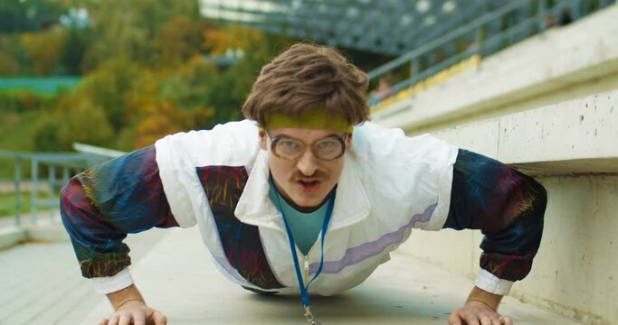 Close up of funny old-fashioned male athlete in big glasses doing push ups outdoors with silly face expression. Caucasian vintage sportsman having workout on street. 70s lifestyle. Sport concept
