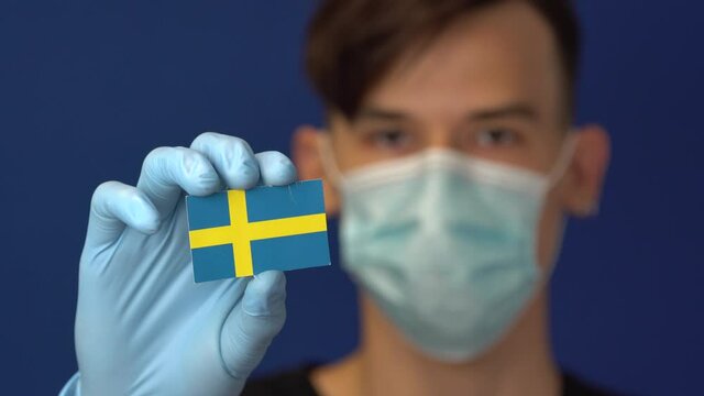 COVID-19 pandemic in Sweden. A young authentic Swede man in protective medical mask and gloves holding the flag of Sweden. The Coronavirus in Europe