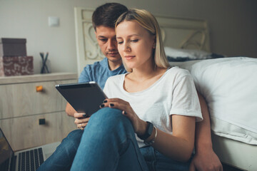 Charming blonde lady and her lover sitting on the floor with a tablet and laptop in the bedroom and spending time together