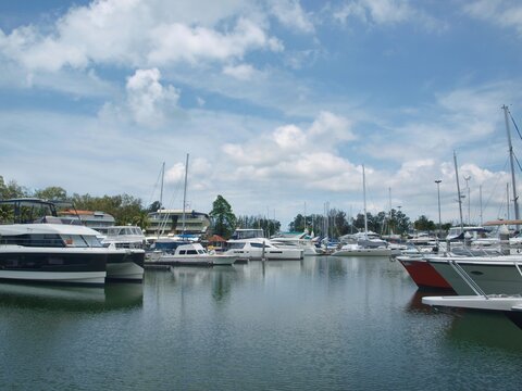 Yachts and boats, mooring location in the harbor. Boating community. Yacht club in harbour, tropical island. Horizontal panoramic view of moored boats and yachts. Water, sky, trees, clouds.