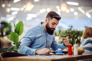 Attractive hungry bearded hipster sitting in restaurant and eating his breakfast.