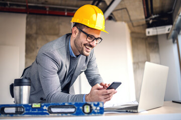 Smiling attractive caucasian architect with helmet on head holding smart phone and looking at laptop while standing in building in construction process. - obrazy, fototapety, plakaty