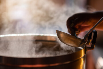 The chef in the restaurant is cooking while using the dipper in a large pot. The water is boiling and the mass of steam reflected in the morning light.