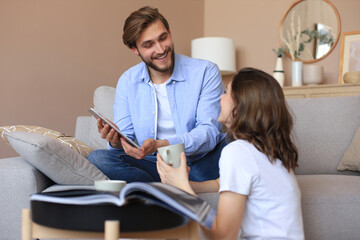 Happy young couple sitting, relaxing on floor in living room watching media content online, doing online shopping in a tablet.