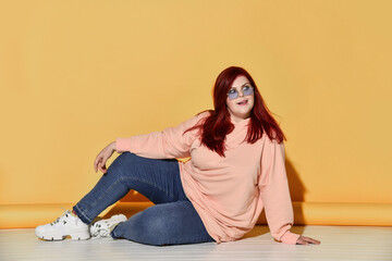Young cheerful red haired overweight woman in casual clothing and sneakers is sitting on floor looking back