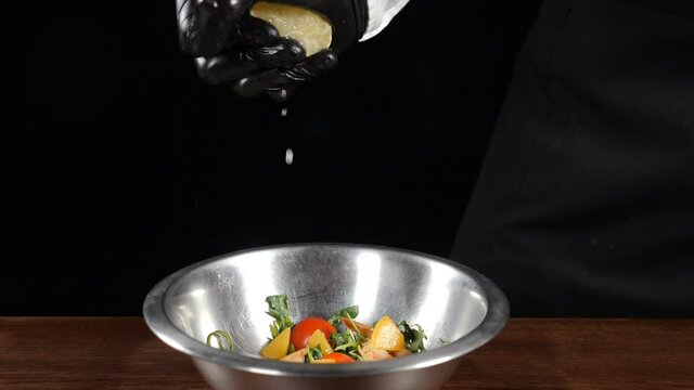 Close-up of chef hand in black gloves, squeezing fresh lemon into steel bowl with fresh salad. Shot on black background. Splashes and drops in slow motion. Full hd