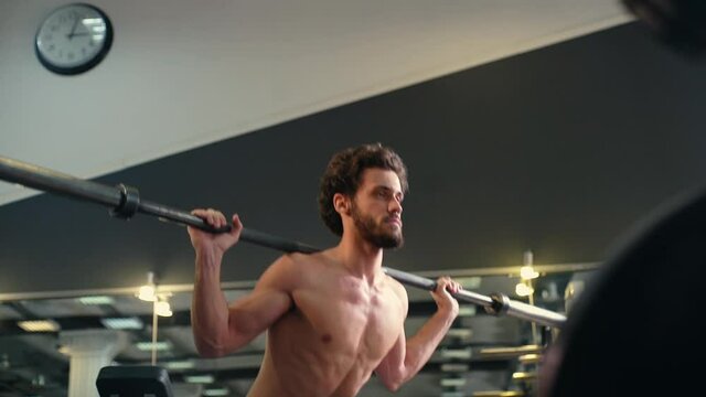 Handsome bearded young man with muscular wiry naked torso squatting using heavy barbell during sport workout training in modern dark gym. Concept of healthy lifestyle. Shooting in slow motion.