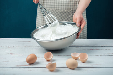 Woman in checkered apron whips egg whites on a white vintage wooden table.