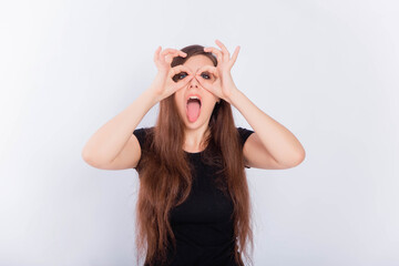 Girl in a black T-shirt with long hair grimaces and shows glasses with her hands isolated on a white background. The concept of an emotional portrait.