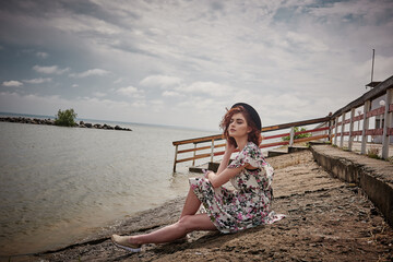 Girl at the sea in a colored dress and hat, sitting on the seashore on a cloudy street