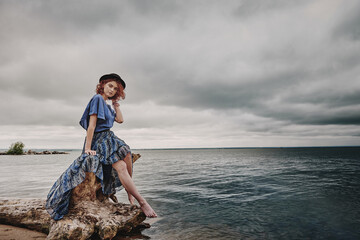 A girl in a blue dress and a black hat sits on the seashore on the street a cloudy sea of dark color.