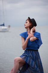 A girl in a blue dress and a black hat sits on the seashore on the street a cloudy sea of dark color.