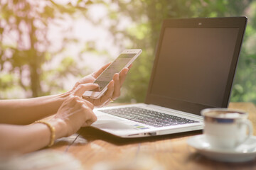 Woman using smartphone with laptop and a cup of coffee on the wooden table outdoor.