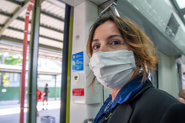 Fototapeta na wymiar Caucasian woman is looking with worried eyes into the camera during her ride with the public transportation train in the city. She wears white medical protection face mask. 
