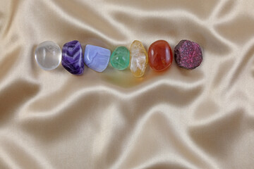 Single row of the Seven chakra healing crystal - magenta, indigo, blue, green, yellow, orange and red coloured gem stones laid on a gold silk material background with copy space below
