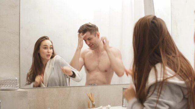 Happy young couple dances and sings in front of bathroom mirror at home.