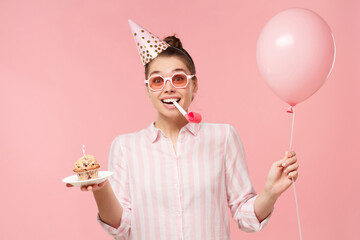 Young happy birthday girl wearing colored glasses and holiday hat, holding balloon, cake and...