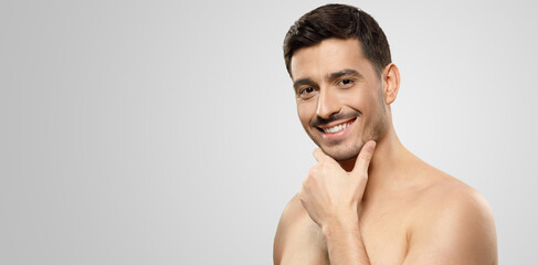 Horizontal banner of young handsome man standing with naked shoukder, holding chin with smile, isolated on gray background with copy space, men beauty ads - obrazy, fototapety, plakaty
