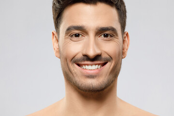 Headshot portrait of handsome man with brown hair and eyes with stubble on face, standing shirtless with happy smile, isolated on gray background