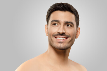 Obraz premium Young handsome man with naked shoulders and bristle, looking aside with dreamful smile, thinking of something pleasant, isolated on gray background