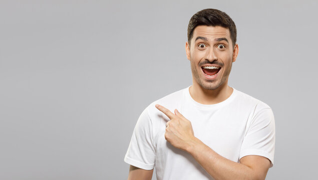 Horizontal Banner Of Young Handsome Man Wearing White T-shirt Pointing Leftwards To Copy Space With Excited Shocked Face, Isolated On Gray Background