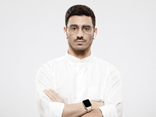 Young handsome man wearing white shirt, glasses, smart watch and wireless earphones, standing with arms crossed, isolated on gray background