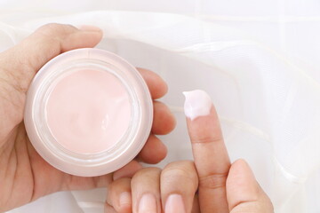 Top view of close up woman finger applying pink cream in jar on white fabric background