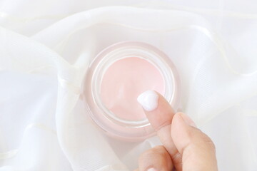 Top view of close up woman finger applying pink cream in jar on white fabric background