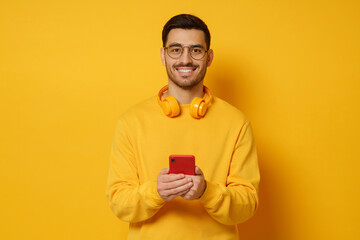 Young man in glasses, looking at camera with happy smile, holding phone in hands, wearing headphones round neck, feeling relaxed, isolated on yellow background