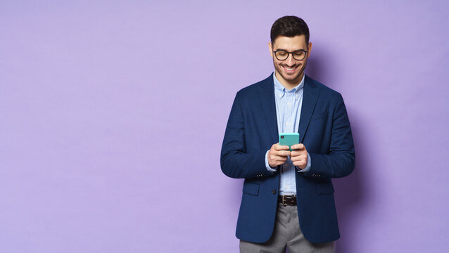Horizontal banner of young business man wearing glasses and formal clothes, texting on phone, smiling happily at screen, isolated on purple background