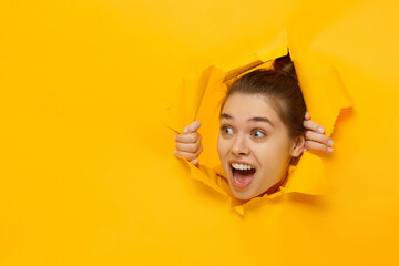 Fototapeta Young excited girl looking through hole in paper at beneficil commercial offer with eyes round with surprise, isolated on yellow background with copy space obraz
