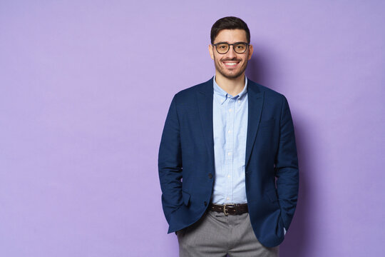 Young businessman wearing formal clothes and eyeglasses, standing isolated against purple background in relaxed pose, smiling friendly, copy space on left