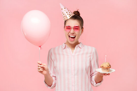 Happy girl in colored eyeglasses, wearing birthday hat, holding balloon and cake with lit candle, laughing with surprise, isolated on pink background