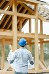 Builder or architect in hard hat supervising a project, standing with blueprints on the construction site. Building wooden frame house concept