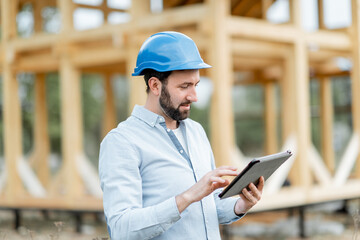 Architect or builder supervising a project with a digital touchpad near the wooden house structure,...